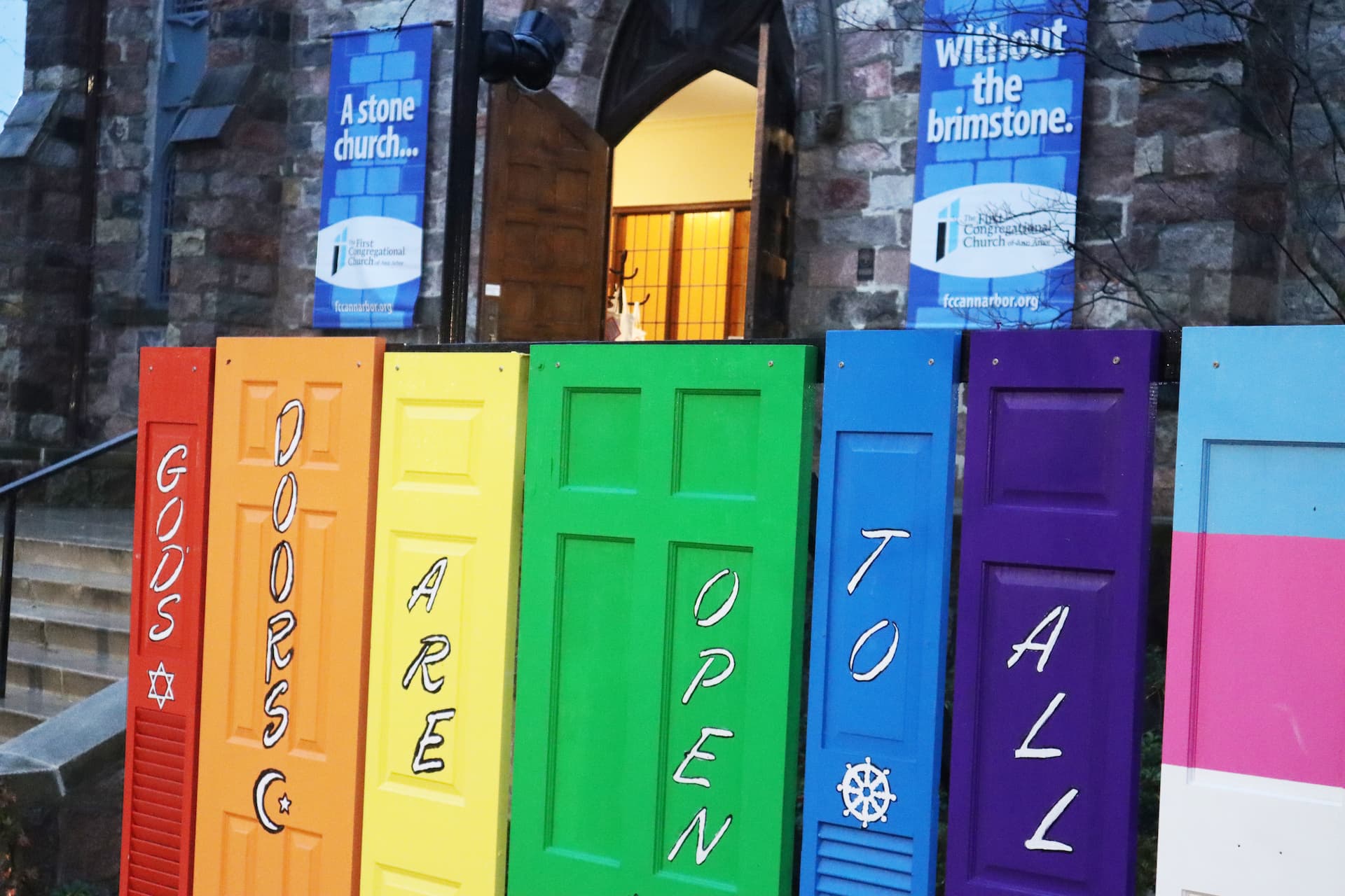 Doors painted to resemble the LGBTQIA+ pride flag with the words God's Doors Are Open To All.